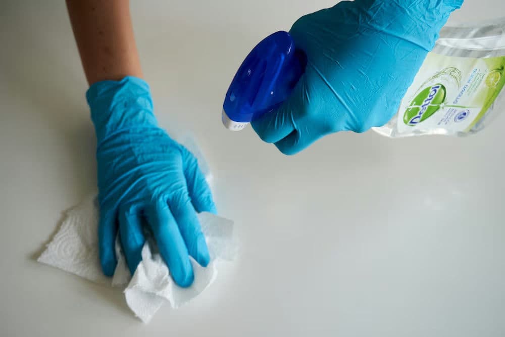 Person in blue gloves cleaning a surface with a spray cleaner and paper towel.